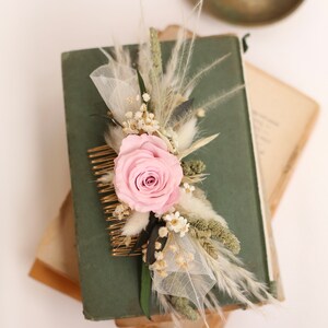 May include: A floral hair comb featuring a light pink rose, dried flowers, and feathery accents. The comb is gold-toned and is set against a dark green book. A vintage-style accessory for weddings or special occasions.