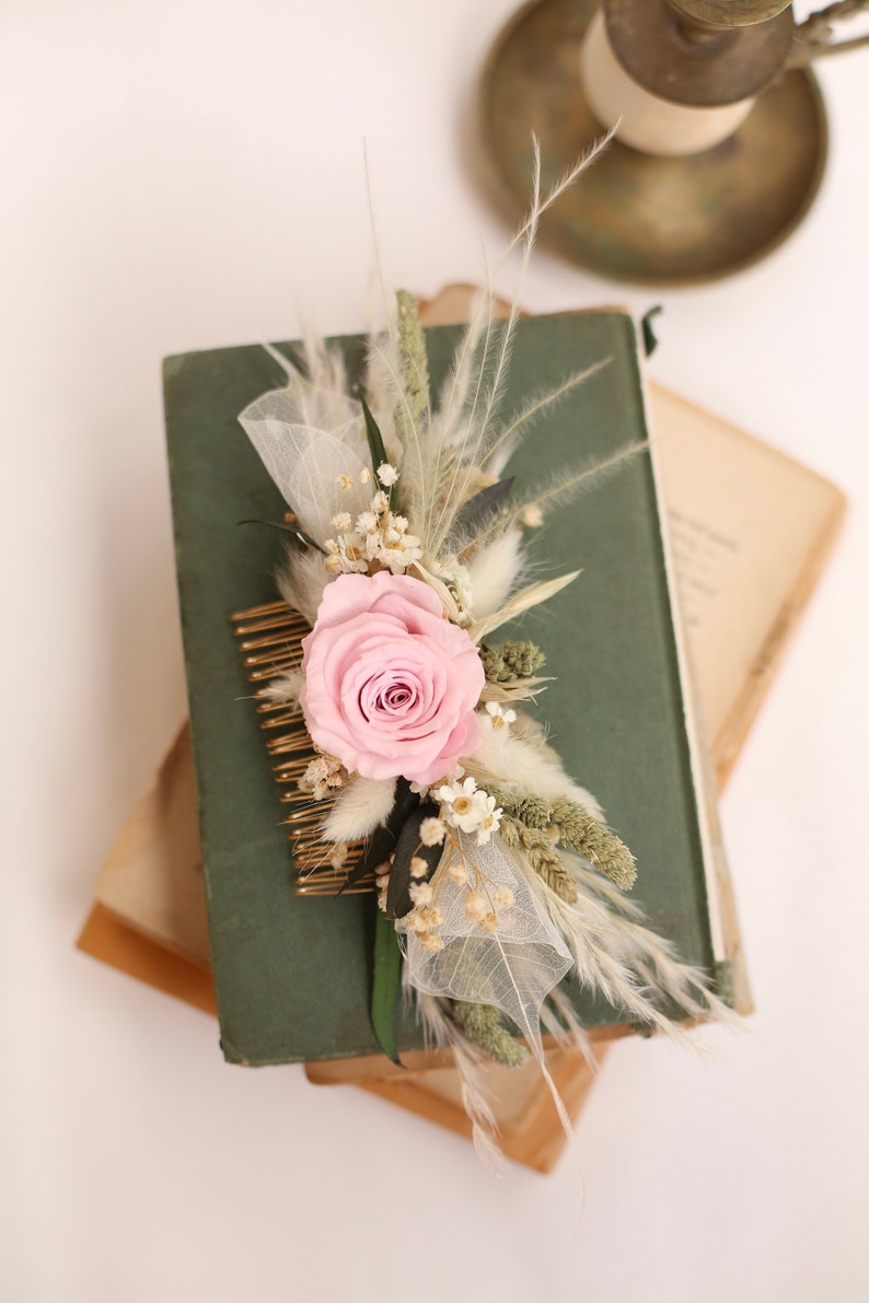 May include: A floral hair comb featuring a light pink rose, dried flowers, and feathery accents. The comb is gold-toned and is set against a dark green book. A vintage-style accessory for weddings or special occasions.