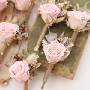 Puede incluir: Cinco boutonnières de rosa rosa con flores secas y vegetación. Cada boutonnière está atada con cuerda y presenta una sola rosa rosa.
