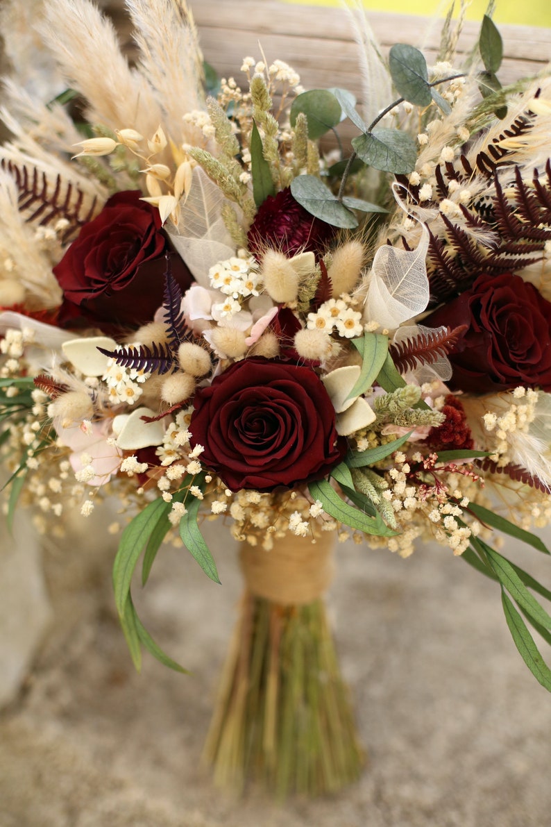 May include: A burgundy and cream dried flower bouquet with burgundy roses, white baby's breath, and dried grasses.