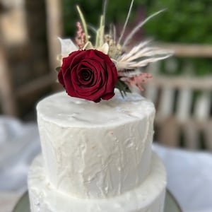 May include: A two-tiered white wedding cake with a single red rose and dried flowers on top. The cake is decorated with a white frosting that has a drippy effect.