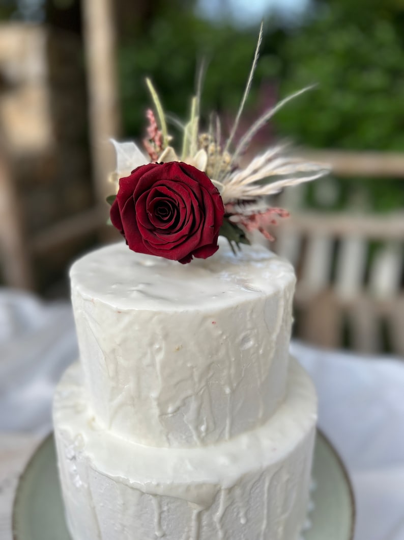 May include: A two-tiered white wedding cake with a single red rose and dried flowers on top. The cake is decorated with a white frosting that has a drippy effect.