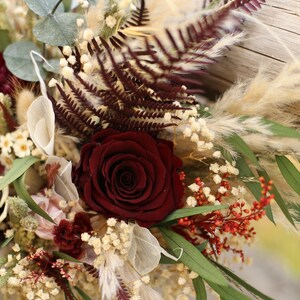 May include: A bouquet of dried flowers with a single dark red rose, white baby's breath, burgundy ferns, and beige pampas grass.