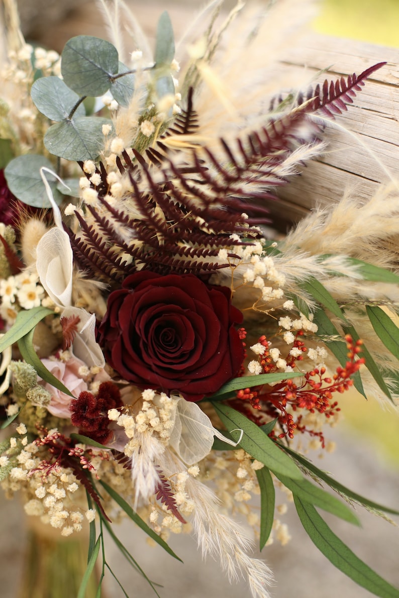 May include: A bouquet of dried flowers with a single dark red rose, white baby's breath, burgundy ferns, and beige pampas grass.