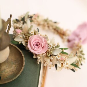 May include: A floral crown featuring a large pink rose as the focal point, surrounded by small white flowers, dried grasses, and green leaves. The crown is set on a green book, with a brass candle holder in the foreground. The overall theme is romantic and natural.