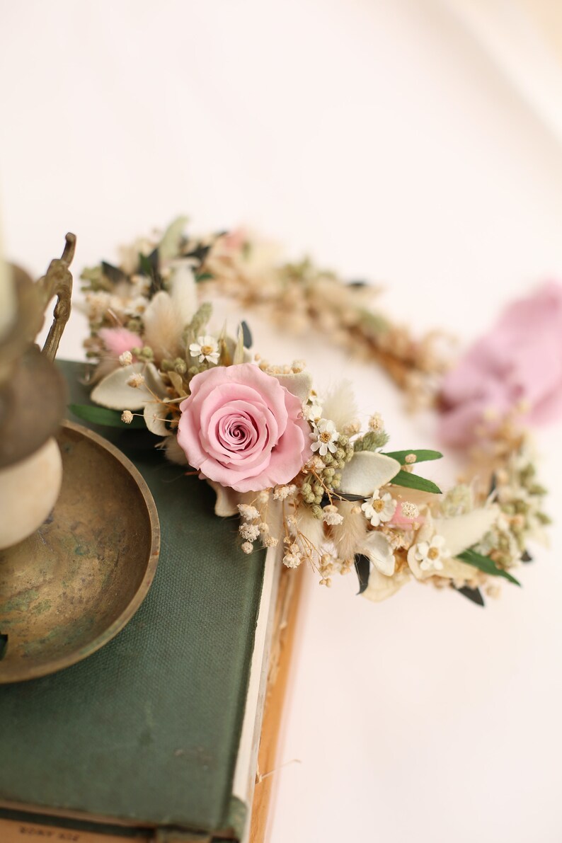 May include: A floral crown featuring a large pink rose as the focal point, surrounded by small white flowers, dried grasses, and green leaves. The crown is set on a green book, with a brass candle holder in the foreground. The overall theme is romantic and natural.