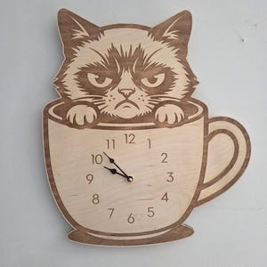 May include: Wooden wall clock featuring a grumpy cat design. The cat is perched inside a coffee cup, with the clock face displaying the time. The clock has a light brown color and is made of wood.
