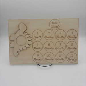May include: A rectangular light-colored wooden milestone board featuring a laser-cut dinosaur shape on the left. The board includes engraved circles labeled Hello World and 1 Month through 12 Months, designed for tracking a baby's growth over time.