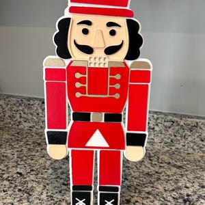 May include: A wooden Christmas nutcracker figurine in red, black, and gold. The figurine has a red hat, jacket, and pants, with black boots and a black belt. The face is tan with black hair and a mustache. The figurine is a festive holiday decoration.