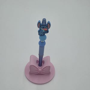 May include: A blue pen featuring a Stitch character topper with two round beads, standing upright in a light pink circular base shaped like a bow. The pen has a metallic blue barrel and the Stitch figure is depicted with its tongue sticking out.