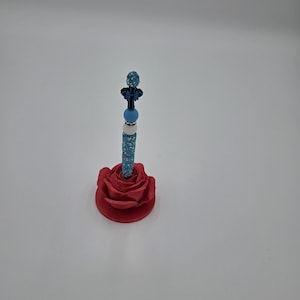 May include: A vertical pen featuring a series of stacked beads in shades of blue, white, and dark blue, topped with a glittery blue sphere. The pen is inserted into a red, sculpted rose-shaped base that serves as a decorative holder.