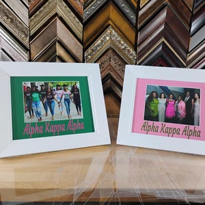 以下が含まれることがあります： ピンクのスクリプトで「Alpha Kappa Alpha」の文字が入った、白いフレームの2枚の写真。1枚は緑色のマットで女性のグループが写っています。もう1枚はピンク色のマットで女性のグループが写っています。