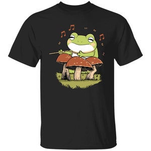 May include: A black t-shirt with a green frog playing the drums on three red and brown mushrooms. The frog is wearing a smile and has closed eyes. The background is black with musical notes.