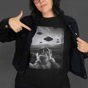 May include: Black t-shirt featuring a black and white image of a monkey taking a selfie with UFOs in the background. The image has a vintage, distressed look. The shirt is being pointed at by a person wearing a black jacket.