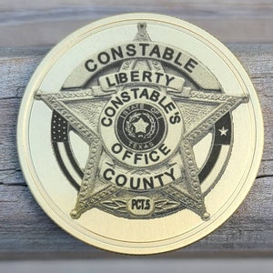 Pode incluir: Um distintivo de metal dourado com uma estrela de cinco pontas no centro. A estrela tem as palavras "Constable's Office County" ao redor. O distintivo também tem as palavras "Constable Liberty" no topo.