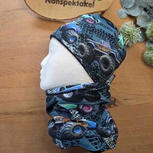 Könnte beinhalten: Blau-schwarzes Mütze- und Halstuch-Set mit Monster-Truck-Design. Die Mütze und das Halstuch sind mit Monstertrucks, Reifenspuren und verschiedenen Farben gemustert. Das Set wird auf einem Schaufensterkopf präsentiert.