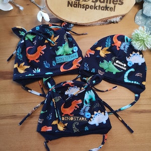 Könnte beinhalten: Drei marineblaue Baby-Mützen mit einem Dinosaurier-Print in Orange, Grün und Weiß. Jede Mütze hat Ohrenklappen und Bindebänder. Eine Mütze hat den Text "DINOSTARK" auf einem Etikett. Die Mützen werden auf einer Holzoberfläche präsentiert.