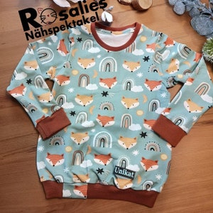 Könnte beinhalten: Ein hellblaues Sweatshirt mit einem sich wiederholenden Muster aus orangefarbenen Füchsen, Regenbogen, Wolken, Sternen und Monden. Die Bündchen, der Halsausschnitt und der Saum sind braun. Der Text "Rosalies Nähspektakel" und "Unikat" sind sichtbar.