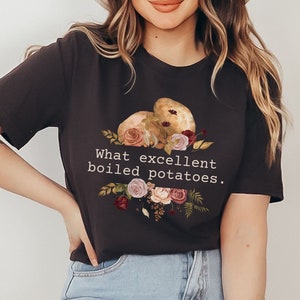 Puede incluir: Una camiseta negra con un gráfico de dos patatas y flores. El texto "What excellent boiled potatoes." está impreso en la camiseta.