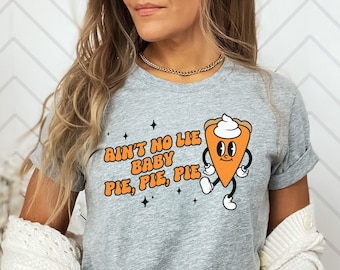 May include: A gray t-shirt with the text "Ain't No Lie Baby Pie, Pie, Pie" and a cartoon pie slice graphic. The pie slice has a whipped cream topping and is walking. An orange pumpkin is held in the hands.
