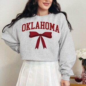May include: Gray sweatshirt with the word "OKLAHOMA" printed in red and a red bow. The sweatshirt is being worn by a person.