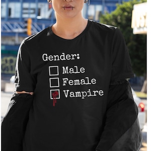 May include: Black t-shirt with a white graphic that says "Gender:" with a checkbox next to each option: "Male", "Female", and "Vampire". The checkbox next to "Vampire" is checked and has a red blood drop dripping from it.