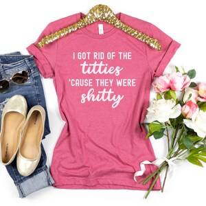 Op de afbeelding: Een roze t-shirt met de tekst "I GOT RID OF THE titties 'CAUSE THEY WERE shitty" in het wit. Het shirt wordt getoond met een blauwe jeans, gouden ballerina's en een zonnebril. Er is ook een boeket bloemen aanwezig.