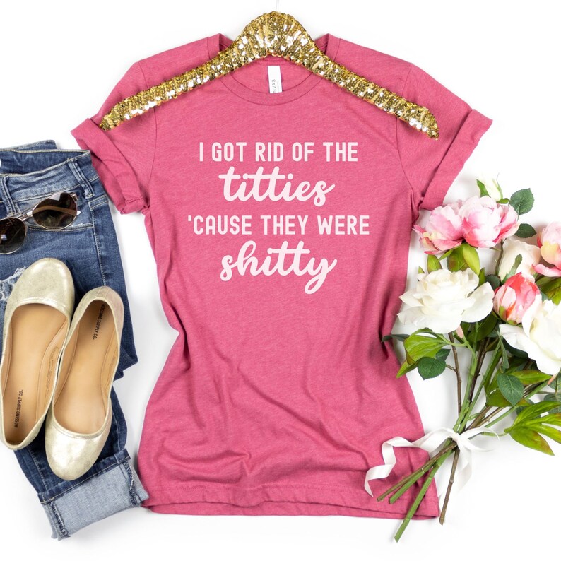 Op de afbeelding: Een roze t-shirt met de tekst "I GOT RID OF THE titties 'CAUSE THEY WERE shitty" in het wit. Het shirt wordt getoond met een blauwe jeans, gouden ballerina's en een zonnebril. Er is ook een boeket bloemen aanwezig.