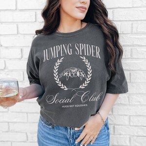 May include: A dark gray t-shirt with a white graphic design that says "Jumping Spider Social Club" with a spider illustration and the text "Hugs not squishes".