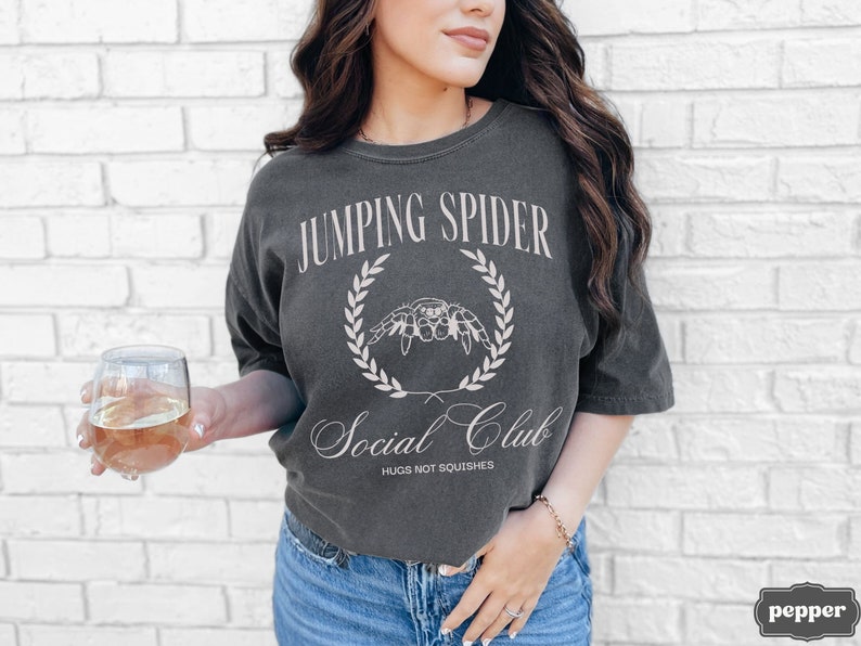 May include: A dark gray t-shirt with a white graphic design that says "Jumping Spider Social Club" with a spider illustration and the text "Hugs not squishes".