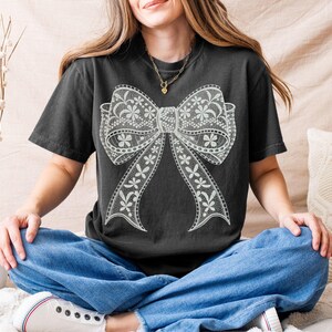 May include: A black t-shirt featuring a large, white lace bow design. The bow has intricate floral patterns and detailed edges. The shirt is a dark charcoal color and has short sleeves.