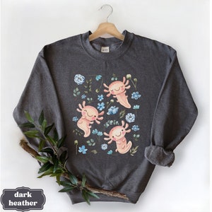 May include: Dark heather grey sweatshirt featuring a print of three pink axolotls swimming in a field of blue and white flowers.