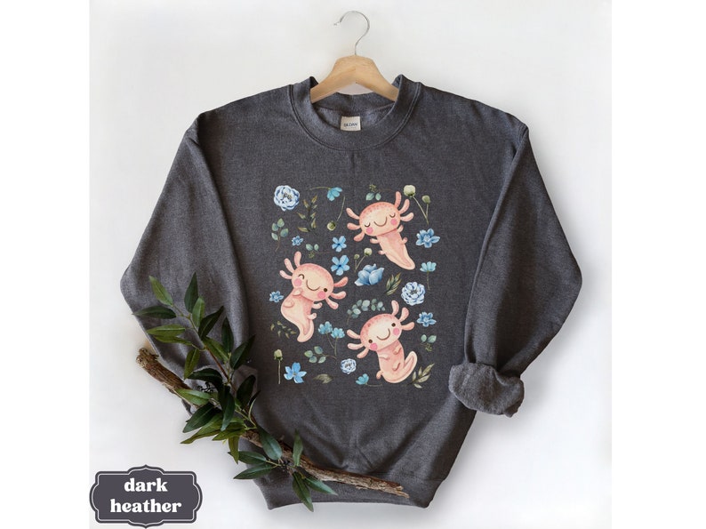 May include: Dark heather grey sweatshirt featuring a print of three pink axolotls swimming in a field of blue and white flowers.