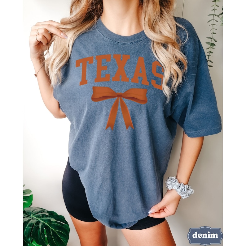 May include: A navy blue t-shirt with the word "TEXAS" printed in orange and a large orange bow underneath.