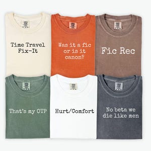 May include: Six t-shirts in various colors, each with a different text design. The shirts are cream, orange, brown, green, white, and gray. The text includes phrases like "Time Travel Fix-It" and "Hurt/Comfort."