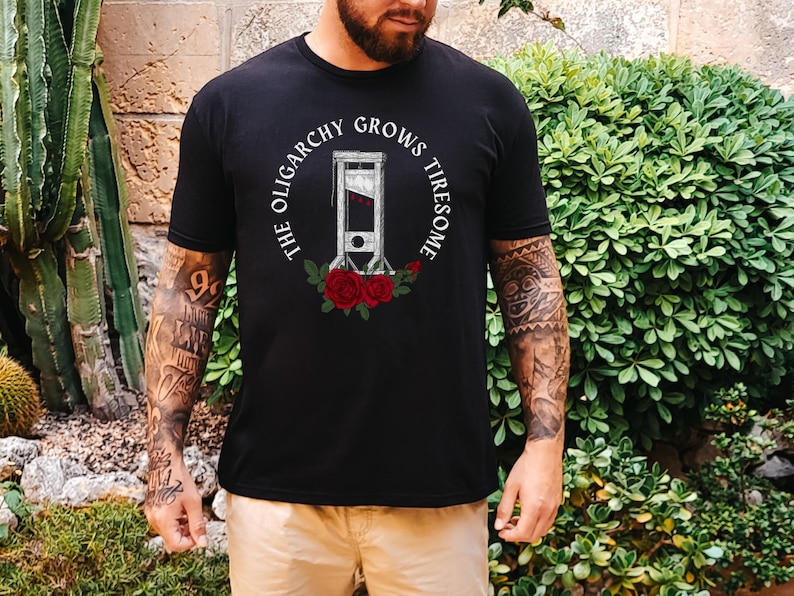 May include: A black t-shirt with a white graphic print of a guillotine with red roses and the text 'The Oligarchy Grows Tiresome'.