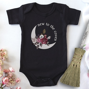 K&ouml;nnte beinhalten: Schwarzer Baby-Strampler mit einem wei&szlig;en Halbmond und einem Blumendesign. Der Text "new to the coven" ist auf dem Strampler aufgedruckt.
