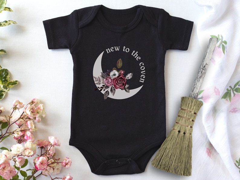 K&ouml;nnte beinhalten: Schwarzer Baby-Strampler mit einem wei&szlig;en Halbmond und einem Blumendesign. Der Text "new to the coven" ist auf dem Strampler aufgedruckt.