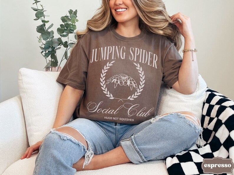 May include: A woman wearing a brown t-shirt with the text "Jumping Spider Social Club Hugs Not Squishes" and a white spider graphic. She is sitting on a white couch with a black and white checkered blanket.