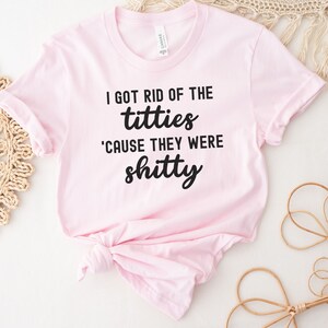 Op de afbeelding: Lichtroze T-shirt met zwarte tekst: "I GOT RID OF THE titties 'CAUSE THEY WERE shitty". Het T-shirt is aan de zoom geknoopt. De stof lijkt zacht en comfortabel.