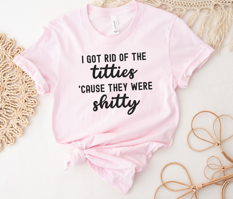 Op de afbeelding: Lichtroze T-shirt met zwarte tekst: "I GOT RID OF THE titties 'CAUSE THEY WERE shitty". Het T-shirt is aan de zoom geknoopt. De stof lijkt zacht en comfortabel.
