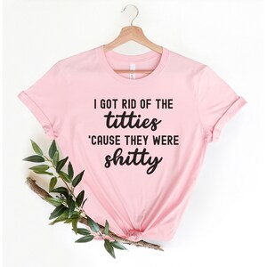 Op de afbeelding: Lichtroze t-shirt met de zwarte tekst "I GOT RID OF THE titties 'CAUSE THEY WERE shitty". Het shirt hangt aan een houten hanger, met een takje groen.