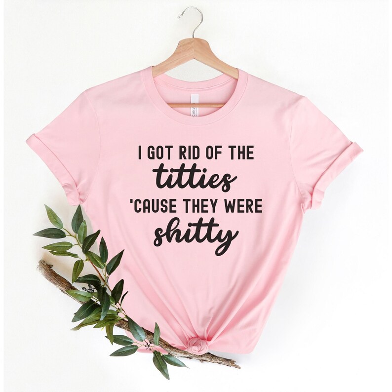 Op de afbeelding: Lichtroze t-shirt met de zwarte tekst "I GOT RID OF THE titties 'CAUSE THEY WERE shitty". Het shirt hangt aan een houten hanger, met een takje groen.