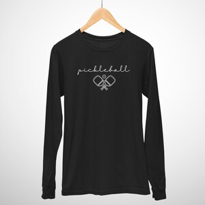 May include: Black long-sleeve shirt with the word "pickleball" in a cursive font. Below the word is a graphic of two crossed pickleball paddles and a ball. The shirt is hanging on a wooden hanger.