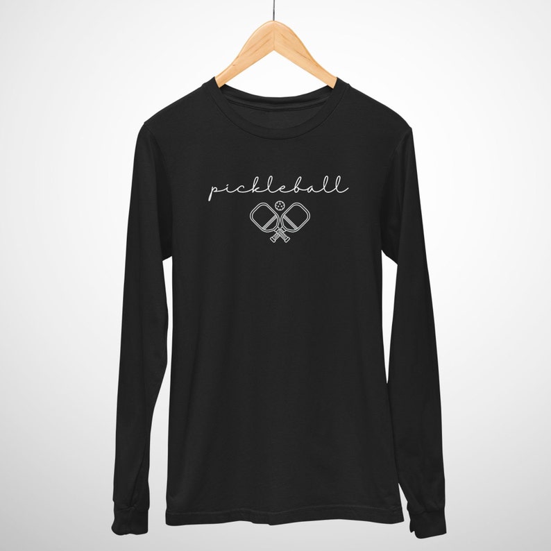 May include: Black long-sleeve shirt with the word "pickleball" in a cursive font. Below the word is a graphic of two crossed pickleball paddles and a ball. The shirt is hanging on a wooden hanger.