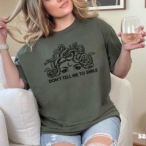 K&ouml;nnte beinhalten: Ein gr&uuml;nes T-Shirt mit einem schwarzen Aufdruck eines Frauenkopfes mit Schlangen als Haaren. Unter dem Aufdruck steht der Text "Don't tell me to smile."