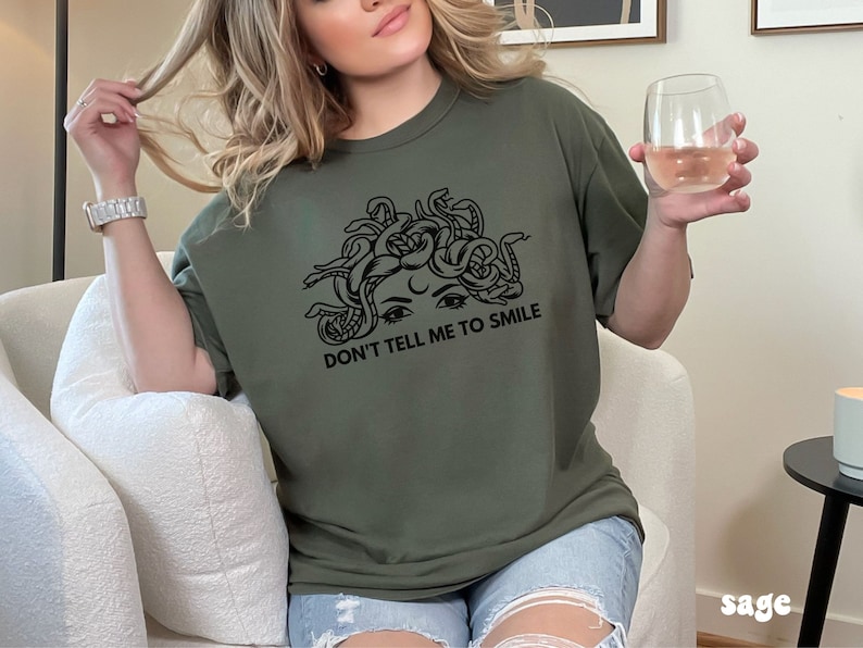 K&ouml;nnte beinhalten: Ein gr&uuml;nes T-Shirt mit einem schwarzen Aufdruck eines Frauenkopfes mit Schlangen als Haaren. Unter dem Aufdruck steht der Text "Don't tell me to smile."