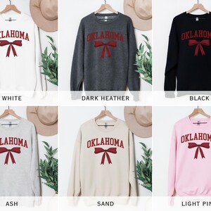 May include: A set of six crewneck sweatshirts with the word "OKLAHOMA" and a red bow printed on the front. The sweatshirts are available in white, dark heather, black, ash, sand, and light pink.