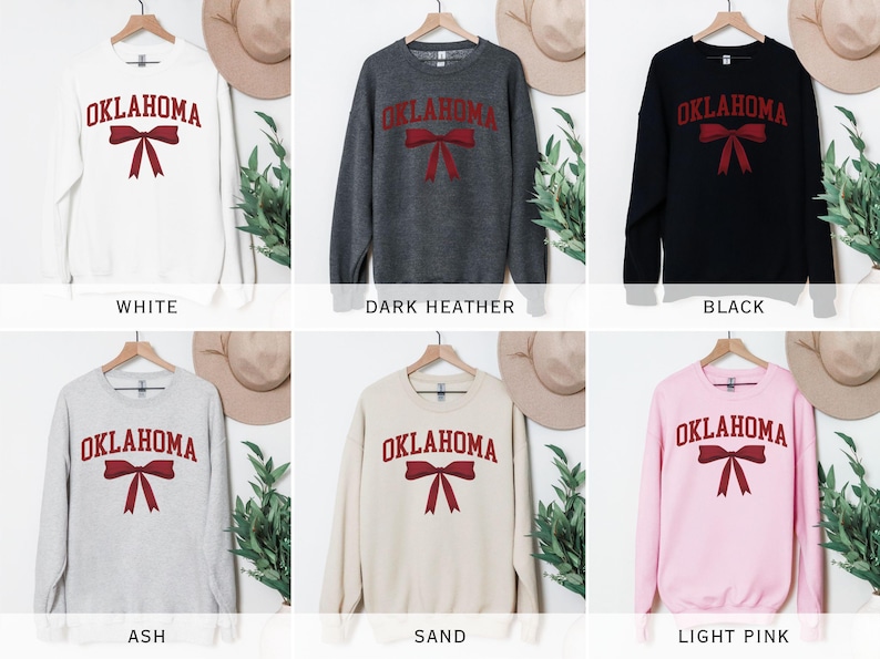 May include: A set of six crewneck sweatshirts with the word "OKLAHOMA" and a red bow printed on the front. The sweatshirts are available in white, dark heather, black, ash, sand, and light pink.