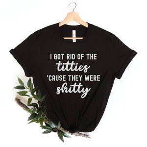 Op de afbeelding: Zwart T-shirt met de witte tekst "I GOT RID OF THE titties 'CAUSE THEY WERE shitty". Het shirt hangt aan een houten hanger tegen een witte achtergrond. Groene bladeren en een tak bevinden zich in de linker benedenhoek.